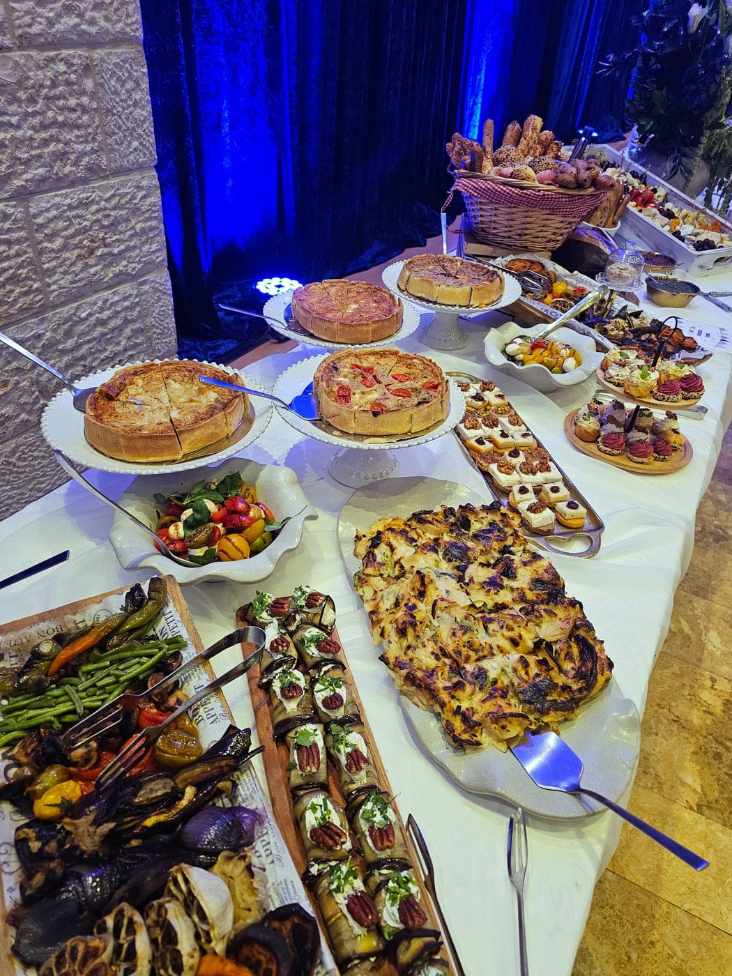 מזנון ארוך בהרמת כוסית — קישים שלמים על סטנדים, פוקצ׳ה בגריל, מגש ירקות צלויים, רולים של חציל וסלסלת מאפים