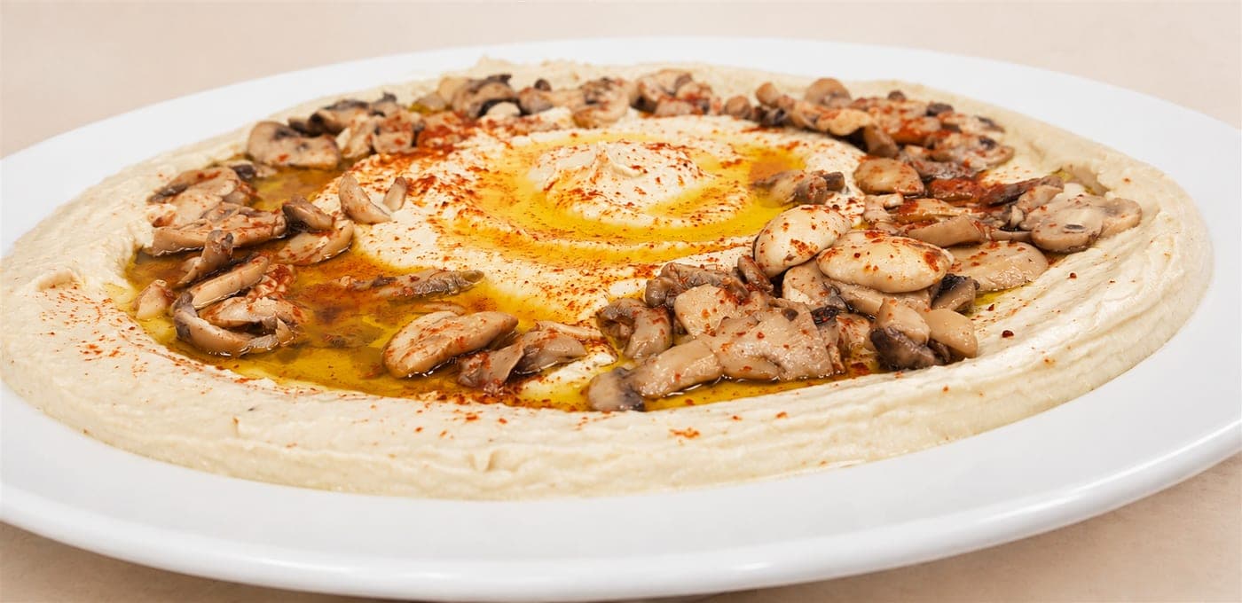 צלחת חומוס לבנה בתקריב — חומוס חלק עם מערבולת רחבה של שמן זית ופפריקה במרכז, ועליו טבעת של פטריות מוקפצות פרוסות בשמן, מתובלות בפפריקה אדומה